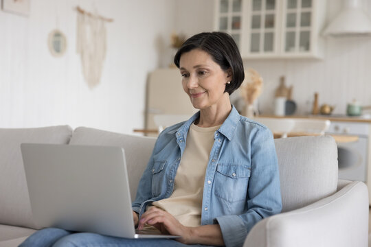 Focused positive senior freelance employee woman working at laptop from home, sitting on couch, typing, smiling, using online application for doing remote job tasks, Internet communication
