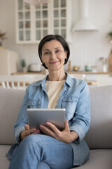 Happy attractive mature freelance professional woman home portrait. Cheerful pretty senior lady in casual holding tablet computer, sitting on sofa, looking at camera, smiling. Vertical shot
