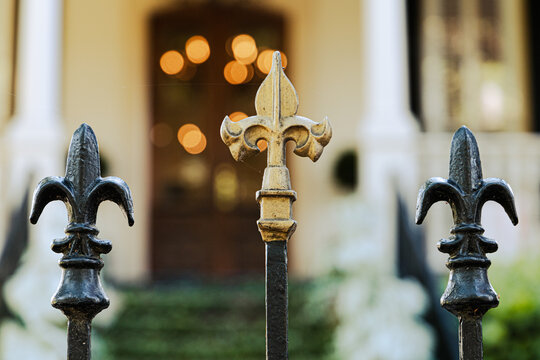 A cast wrought iron fence lined with black and gold fleur de lis post toppers with a New Orleans southern style home in the background