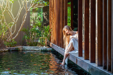 Slow motion footage of woman in white dress relaxing at luxury spa with fish pond. Sitting on wooden bridge and enjoying nature. Zen, meditation.