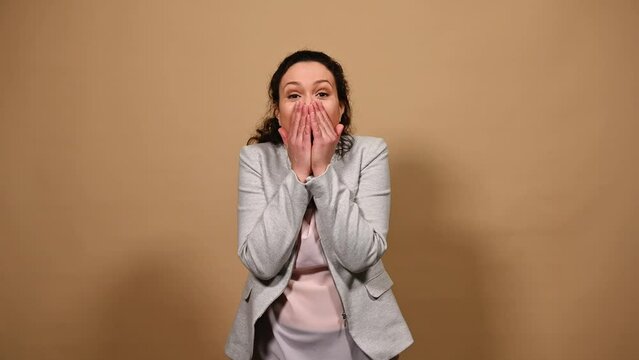 Pretty woman attentively looking into the distance, rejoicing, expressing happiness and positive emotions, keeping her hands palms together, looking at camera with hopeful look on beige background