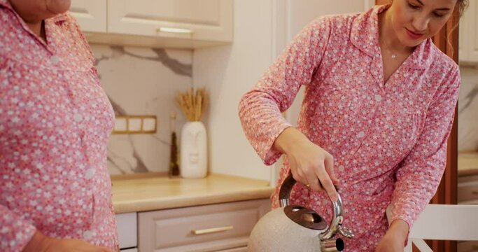 Woman is pouring boiled water to teapot to make tea for her granny in kitchen at home. Tea with homemade bakery. They are wearing in pink pajamas in family look style. Breakfast together in weekend. 