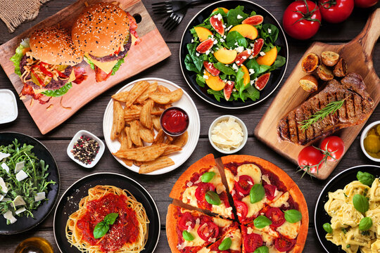 Table scene with assorted delicious foods. Top down view over a dark wood background.