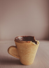 Bitten edible cup made of cookies on a light background. Edible dishes. Cup made of cookies. Bitten cookies in the form of a cup. Cup of coffee. An unusual thing. There is no part of something. 