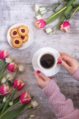 Female hands with a cup of coffee surrounded by the first spring flowers and a plate of cookies on a gray background. Vertical photo. Spring concept. Flat lay. Top view.