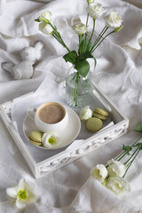 Spring romantic breakfast in bed. A cup of cappuccino, pistachio macaroons and a vase of flowers on a white wooden tray and textile background.  Concept for wishes of a good spring morning.