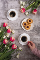 Two cups of freshly brewed coffee, a plate of cookies and tulips with eustoma on a light gray background. Female hand with a cup of coffee surrounded by the first spring flowers on a gray background.