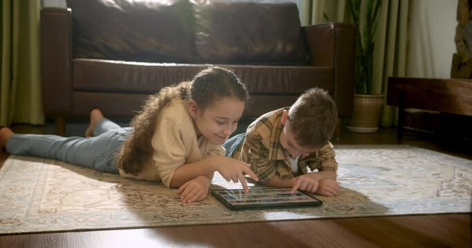 Happy cute two children brother and sister using digital tablet technology device lying on carpet floor, children watch video using a digital tablet. Children having fun in living room at leisure. 4K
