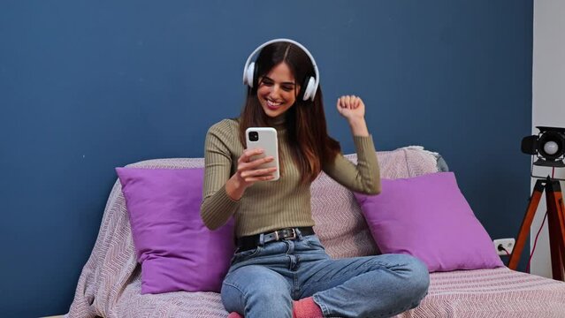Excited young woman listening music with headphones and ajoying dancing on the couch at home using a cellphone app. Slow motion. High quality 4k footage