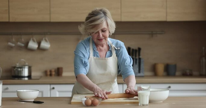 Happy engaged senior baker woman rolling dough, showing bakery food ingredient pitta bread, circular cake ingredient at camera, enjoying cooking, culinary hobby at home kitchen