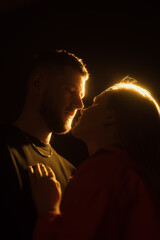 Side view of young bearded man and blond haired woman holding hands and kissing gently in darkness against glowing light