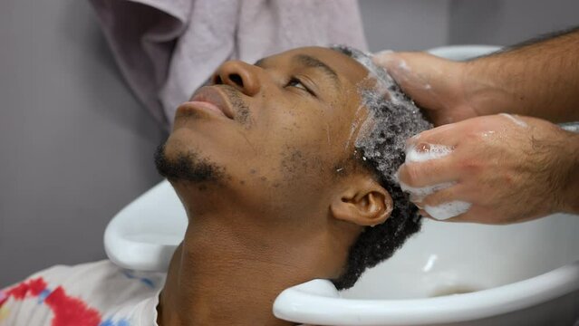 Barber man is washing client afroamerican man hair in barbershop over the sink.