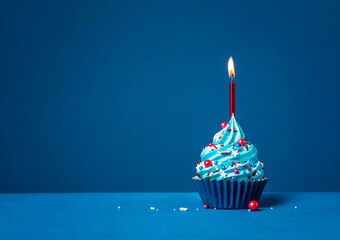 Blue cupcake with red sprinkles and lit candle on a blue background. - 580157809