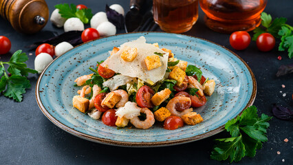 Italian lunch Caesar salad with shrimp, parmesan cheese, lettuce, tomatoes, croutons on a dark table.