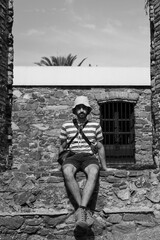 Black and white photo of a young Latin man sitting on a stone wall with a bucket hat looking at the camera. Vertical photo.