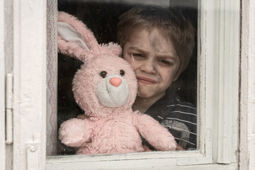 Little grimy boy looking through a dirty old window. A sad smile on his face. He is holding a pink plush rabbit (bunny) in his hands