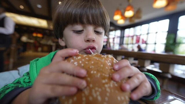 Young boy eating hamburger meal. Child taking a bite of burger food. Kid eats delicious lunch at restaurant diner