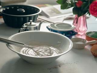 A bowl of white cream with whisk on a table next to a vase of flowers.