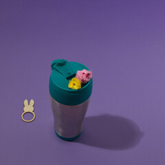 A coffee thermos with little chicks coming out, below is a ring with a rabbit's head and ears, with copy space on a purple background. Minimal Easter scene.