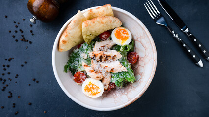 Caesar salad with chicken, parmesan cheese, lettuce, tomatoes, cut tortilla on a dark table.