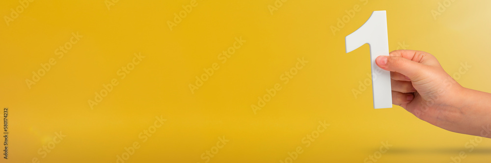 Poster Number one in hand. A hand holds a white number one on a yellow background with copy space. Concept with number one. 1 percent rate, birthday, first or winner.