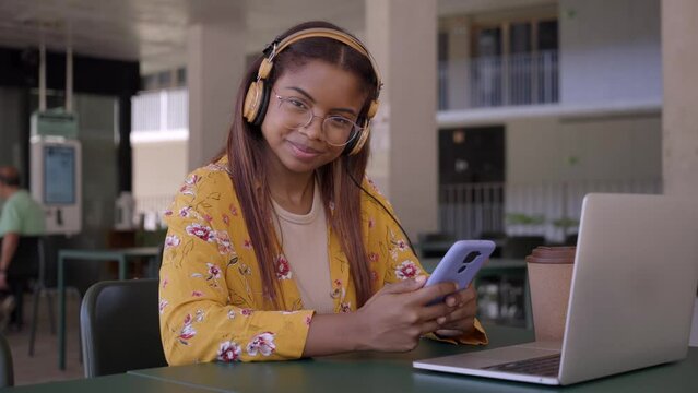Video young black African college student learning online using cell phone. Smiling girl watching virtual education distance learning class studying sitting in campus study room. People looking camera