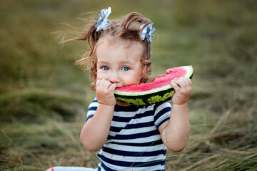 A little pretty girl with two ponytails and wearing a striped t-shirt is eating watermelon.