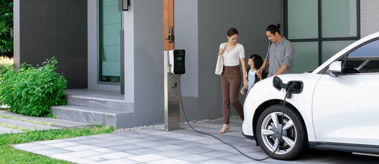 Progressive young parents and daughter with electric vehicle and home charging station. Green and...