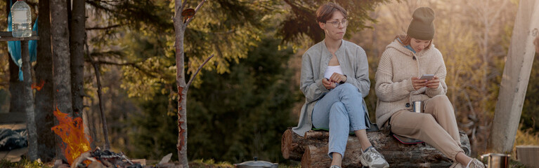 Two young Caucasian women sitting at fire in forest and resting after hiking. Females tourists.