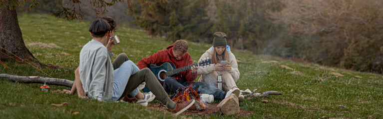 Caucasian young friends sitting at fire in mountains, playing on guitar and singing. Tourism.