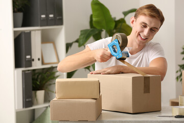 Young man packing box with adhesive tape indoors
