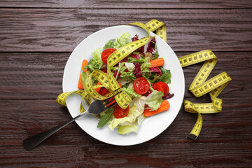 Plate with fresh vegetable salad, fork and measuring tape on wooden table, flat lay. Healthy diet concept
