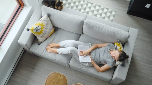 Overhead view of woman reading the book, lying on couch at home