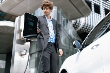 Below view of progressive businessman with electric car recharging at public charging station at...