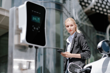 Businesswoman wearing black suit using smartphone, leaning on electric car recharge battery at...