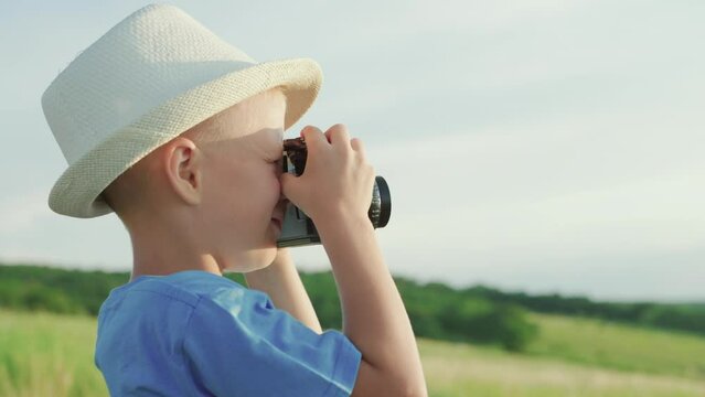 Cute little kid is holding vintage camera and taking pictures of nature. Happy boy dreams of becoming photographer. Kid plays in park in summer, spring. Child dreams of traveling, making discoveries