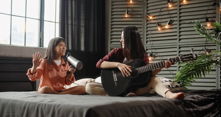 Two asian sisters sitting on bed, elder playing guitar while little kid is singing into hairdryer....