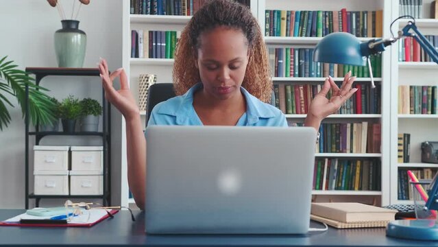 Young progressive African American woman begins to do breathing exercises and meditate like Buddhist yogis to relax between work sits at table with laptop in home office. Businesswoman, freelancer