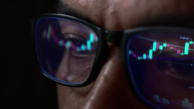 Closeup - Late night eyes of man in spectacles scrolling in front of coder laptop, he is analyzing Bitcoin price chart on cryptocurrency exchange in recession