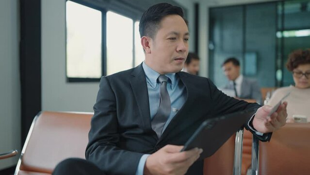 Asian businessman sitting and recheck boarding pass waiting for flight using lablet typing and working at airport terminal