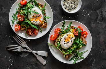 Served breakfast for two - whole grain bread sandwiches with avocado, poached egg and arugula cherry tomatoes salad on a dark background