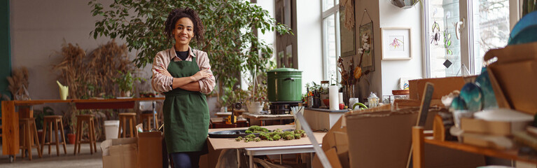 Attractive smiling florist stands with crossed arms on background of her own floral decor studio