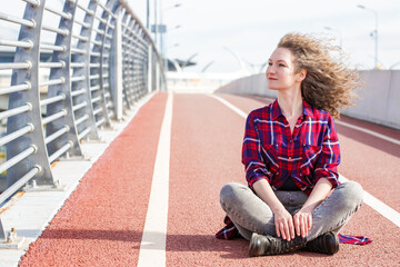 Tired happy woman resting after running, sitting on treadmill and smiling in wind. Summer time. Young athletic woman resting after workout in casual wear with wavy hair. Copy space