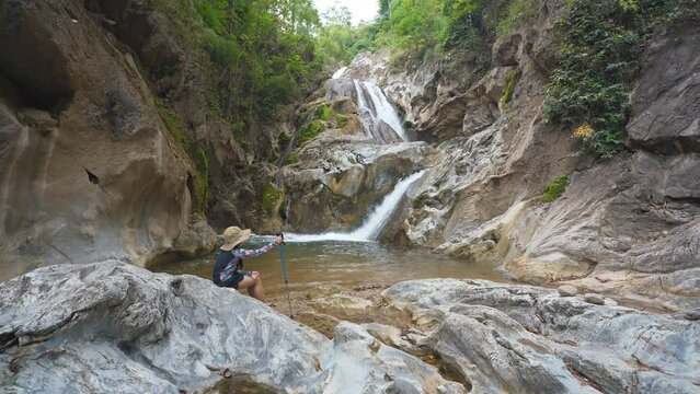 asian child camper or kid girl trekking or hiking by walking pole and wearing hat to sitting on rocks for relax on nature waterfall in forest for holiday adventure travel at lan sang national park