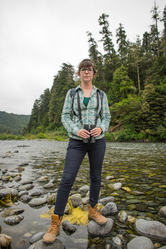 Portrait of female hiker holding binoculars while standing on rocks over stream at Redwood National and State Parks