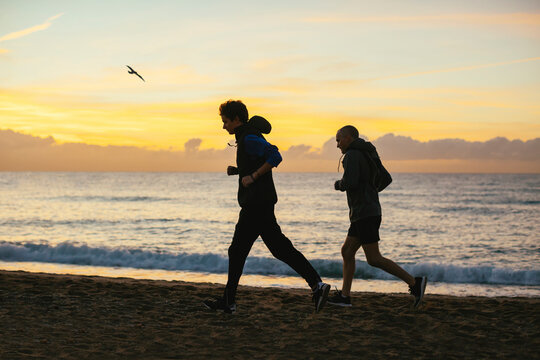 Side view of father and son jogging at beach against cloudy sky during sunset