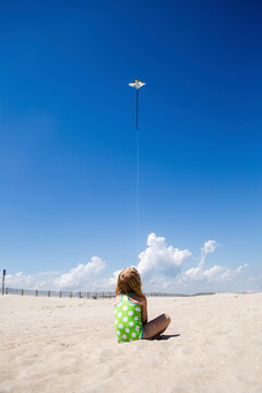 Rear view of girl flying kite while sitting at beach during sunny day