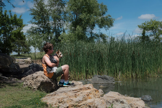 Side view of boy with Yorkshire Terrier sitting on rock by pond during sunny day