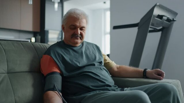 Grandfather Is Checking Blood Pressure, Sitting On Couch, Using Digital Blood Pressure Monitor