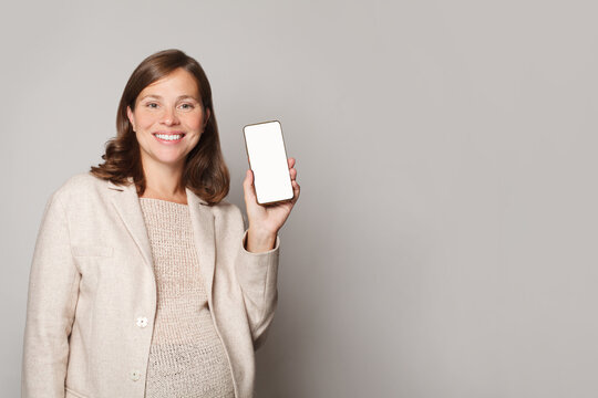 Happy Beautiful Pregnant Woman Showing Her Mobile Phone With White Empty Display And Smiling Against  Grey Studio Wall Background